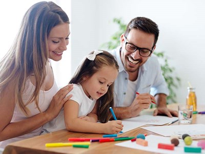 Parents sitting with young child at table smiling while she draws.