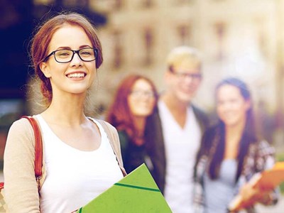 Student smiling with a backpack on and holding a folder in the foreground in front of a building, with other students in the background behind her.