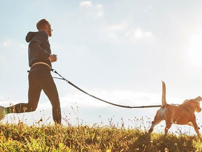 Someone running in a grassy area with their dog on a leash, sunny sky in the background.